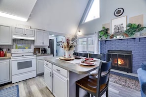 Une cuisine moderne avec des meubles blancs, un îlot central, une cheminée et un coin repas offrant une vue sur l’extérieur.