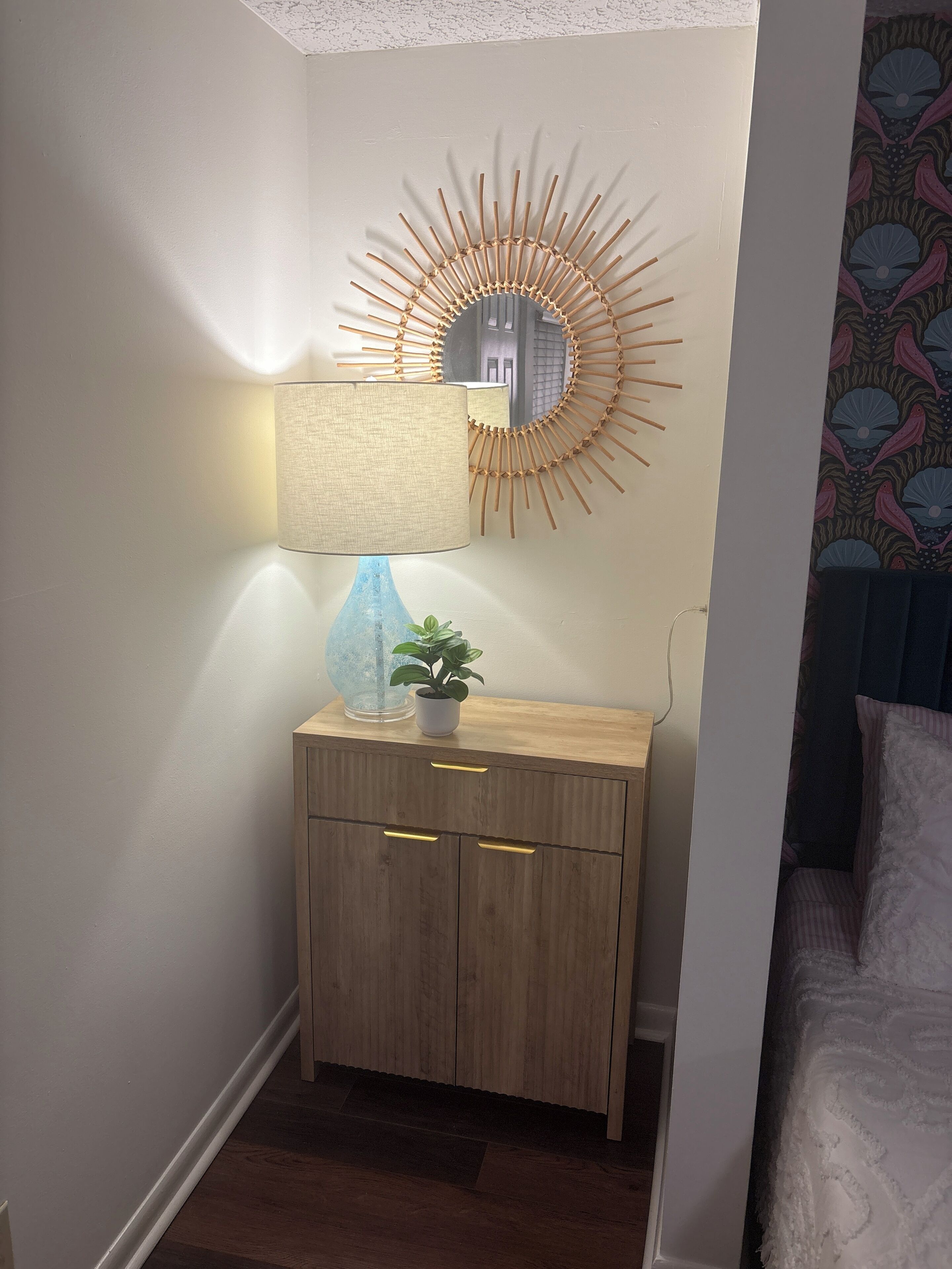 A corner room with a wooden cabinet, a mirror with a sunburst design, a lamp, and a potted plant.