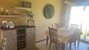 Un coin repas comprenant une table, des chaises, une horloge, une plante et un meuble contenant des bouteilles de vin.