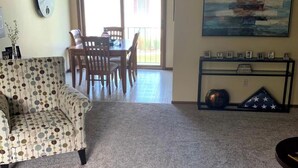 Un salon avec un fauteuil à motifs, une table à manger avec des chaises, un ventilateur de plafond, un grand tableau abstrait et une console sur laquelle sont posées des photos encadrées.
