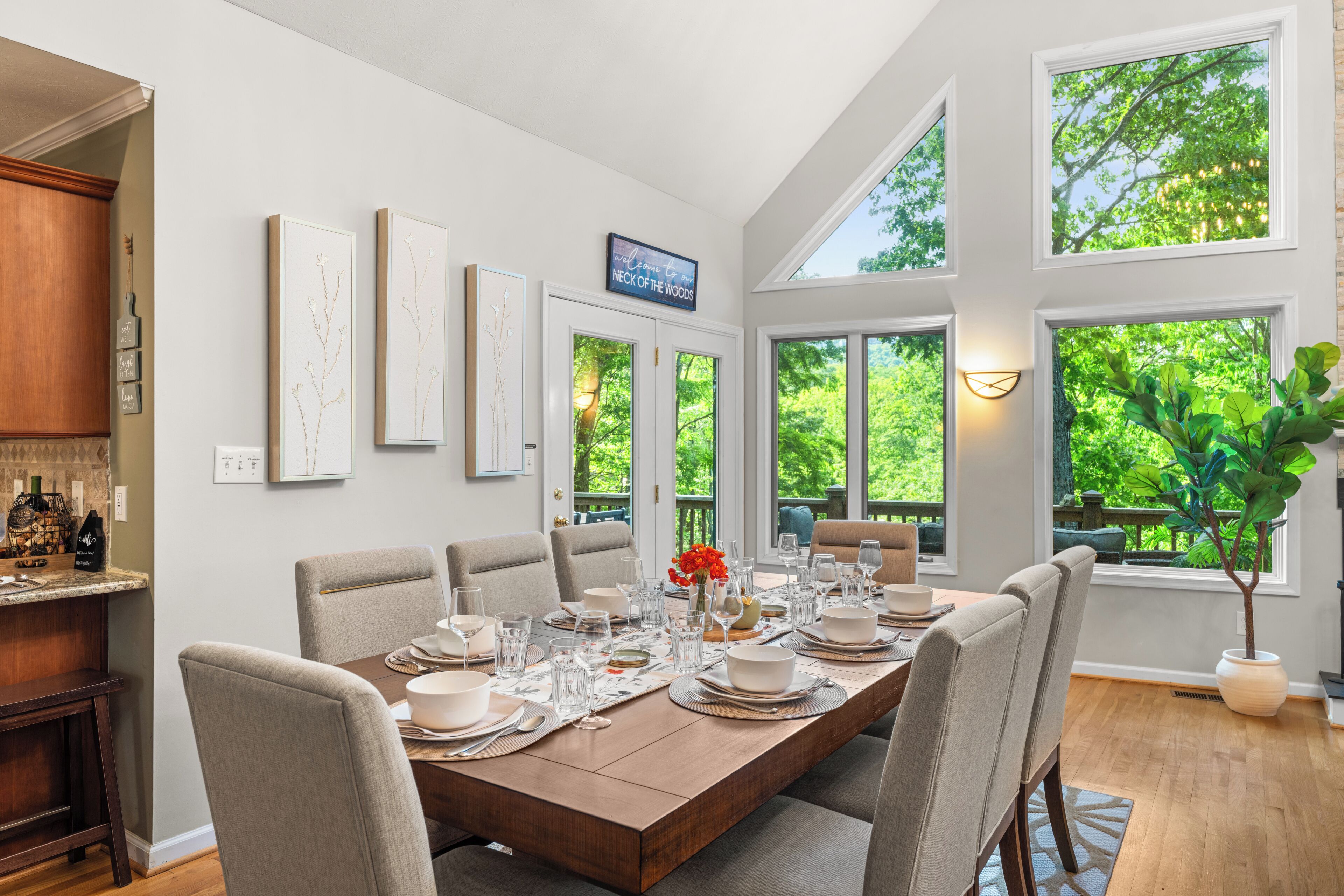 A dining area with a long table set for six, surrounded by chairs, with a view of greenery through large windows.