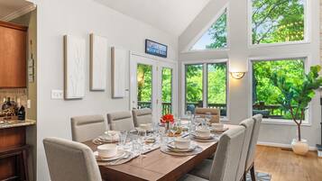 A dining area with a long table set for six, surrounded by chairs, with a view of greenery through large windows.