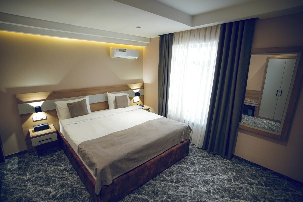 Hotelkamer met een groot bed, nachtkastjes, een airconditioner, een spiegel en een raam met gordijnen.