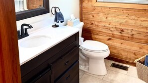 A bathroom with a wooden wall, a white sink, a black faucet, a toilet, a shelf with a plant, and a framed picture. - Lakefront Rustic 4-Bedroom House with Private Dock (Lake Ozark)