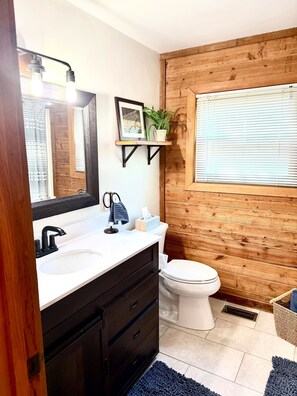 Un baño con pared de madera, un lavabo blanco, un grifo negro, un inodoro, una estantería con una planta y un cuadro enmarcado.