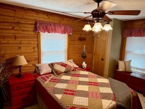 A bedroom with a wooden wall, a bed with a checkered quilt, a red nightstand, a lamp, and a ceiling fan.