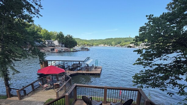 A wooden deck with chairs and a table, overlooking a lake with docks and boats. - Lakefront Rustic 4-Bedroom House with Private Dock (Lake Ozark)