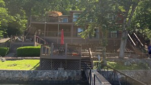 A wooden deck over a body of water, a multi-level building with outdoor seating, and a stone wall. - Lakefront Rustic 4-Bedroom House with Private Dock (Lake Ozark)