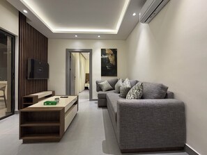 A modern living room with a grey sofa, a wooden coffee table, a flat-screen TV, and a ceiling-mounted air conditioner. - 005 JRE Apartment With Stunning Boulevard Veiw (Amman)
