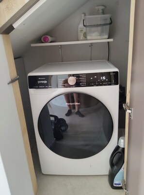A compact, white, front-loading washing machine with a digital display, situated in a small laundry room with a shelf above containing a bottle and a container.