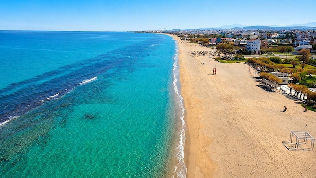 A coastal beach with clear turquoise waters, a sandy shore, and a row of buildings along the coastline. - Fems Luxury 3-level Home w Private Jacuzzi (Heraklion)