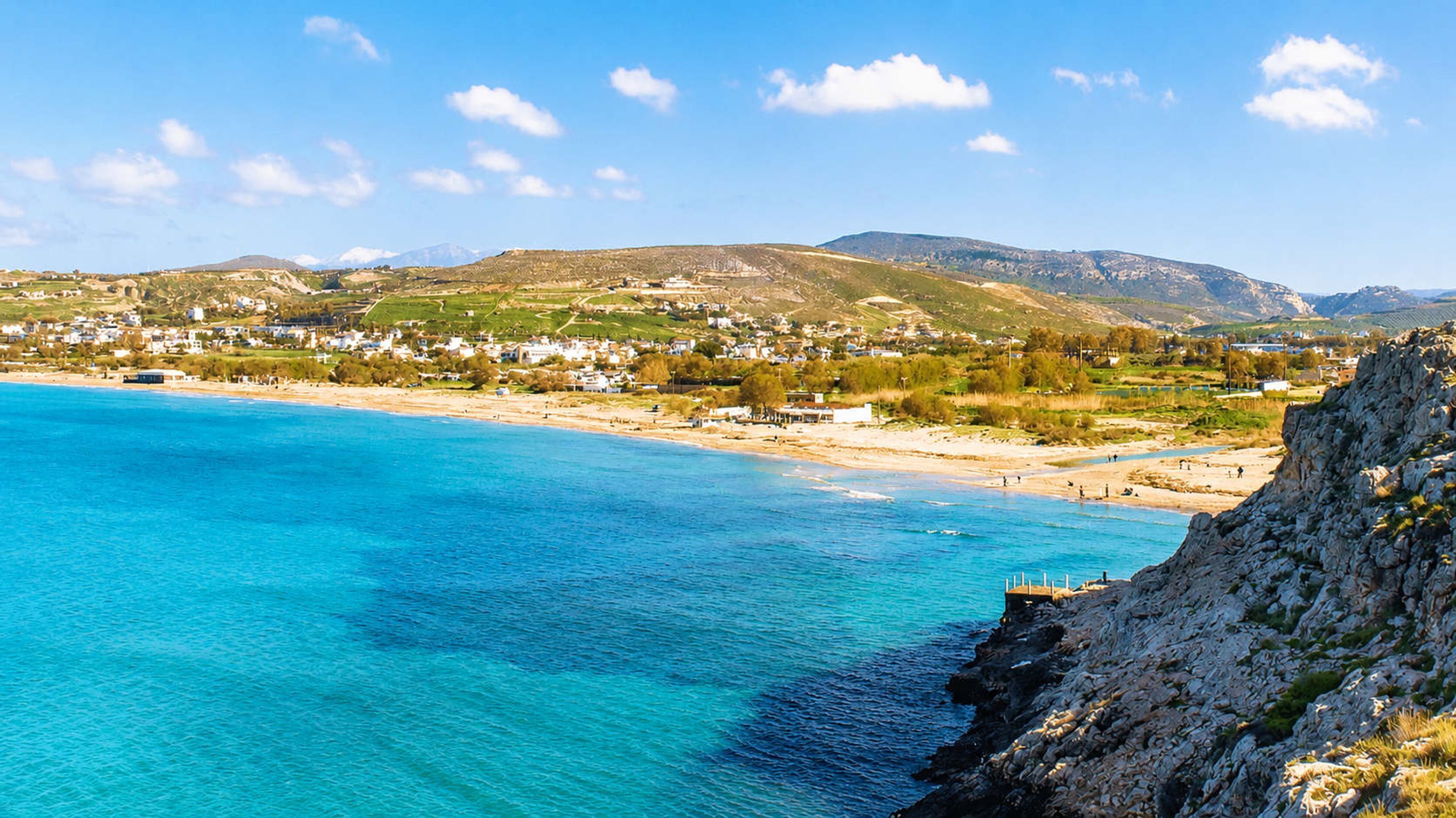 A coastal town with a clear blue sea, rocky coastline, and a hilly backdrop.