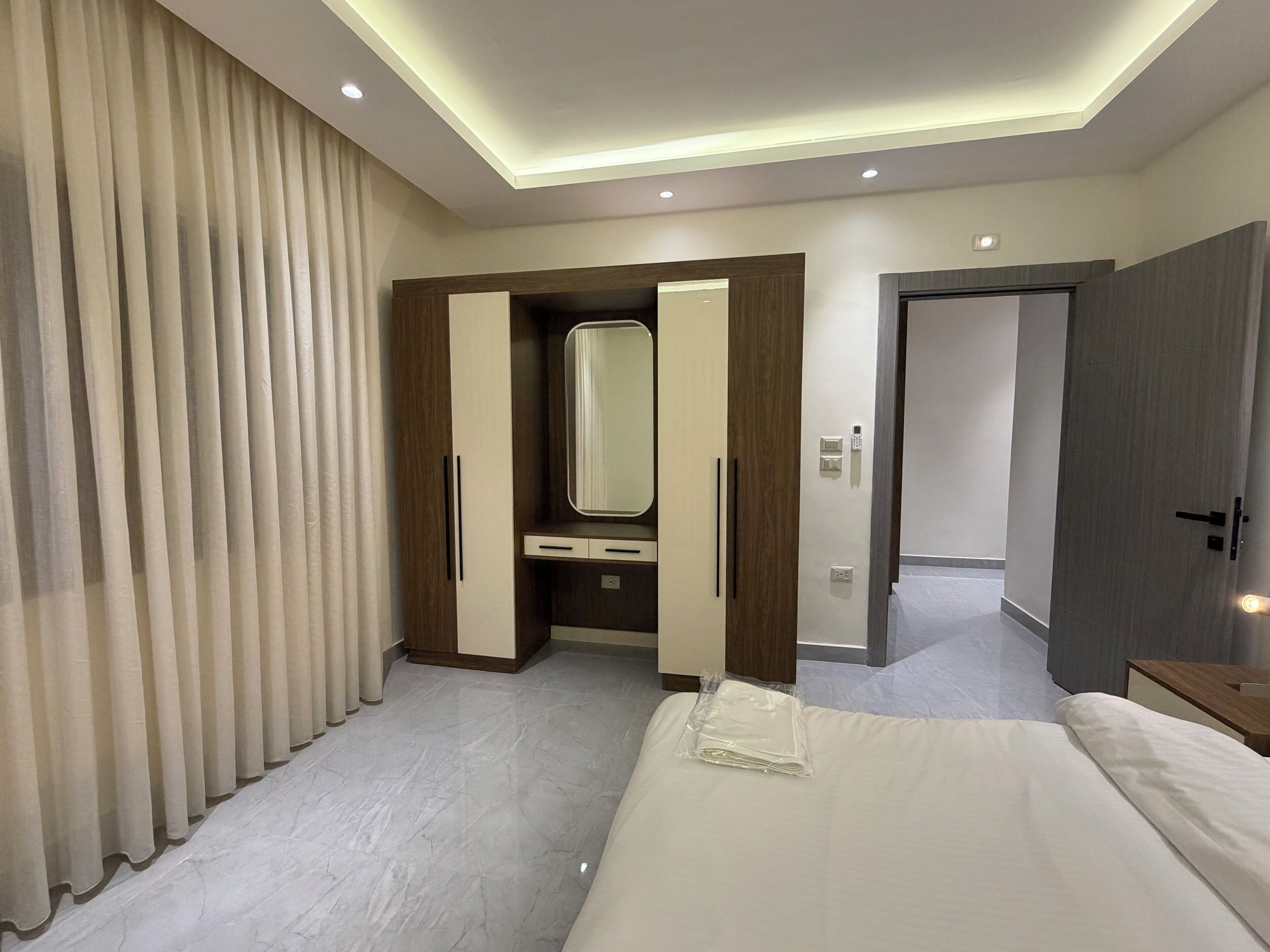 A modern hotel room with a bed, a vanity with a mirror, a door, and a lighted ceiling.