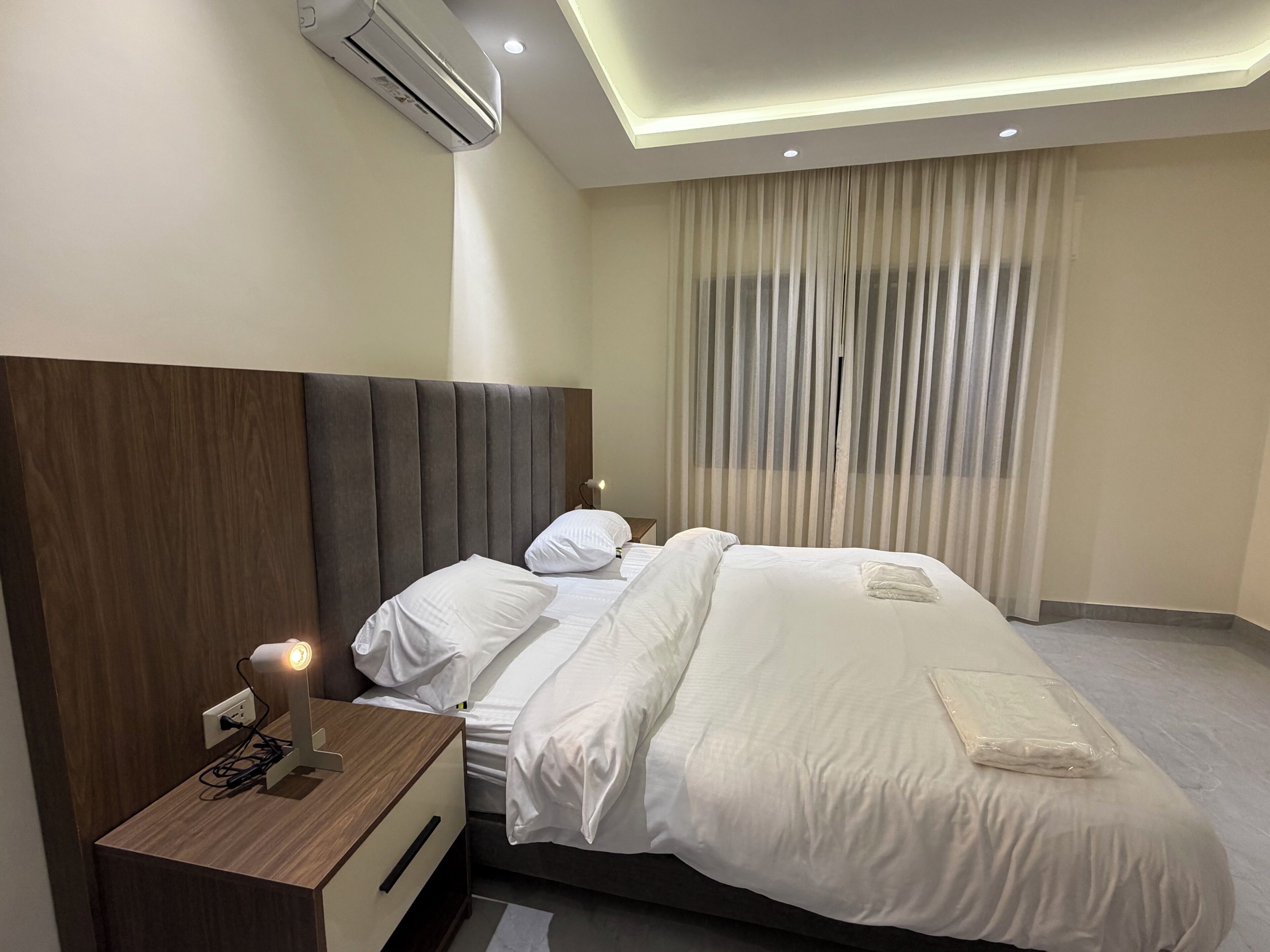 Una habitación de hotel con una cama grande, una mesita de noche con lámpara, aire acondicionado y una ventana con cortinas.