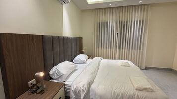 Una habitación de hotel con una cama grande, una mesita de noche con lámpara, aire acondicionado y una ventana con cortinas.