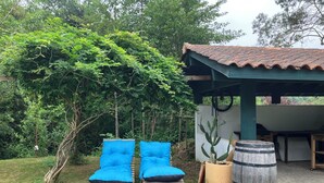 A covered outdoor seating area with two blue cushioned chairs, a cactus, and a wooden barrel. - Friendly house surrounded by greenery. (Biriatou)