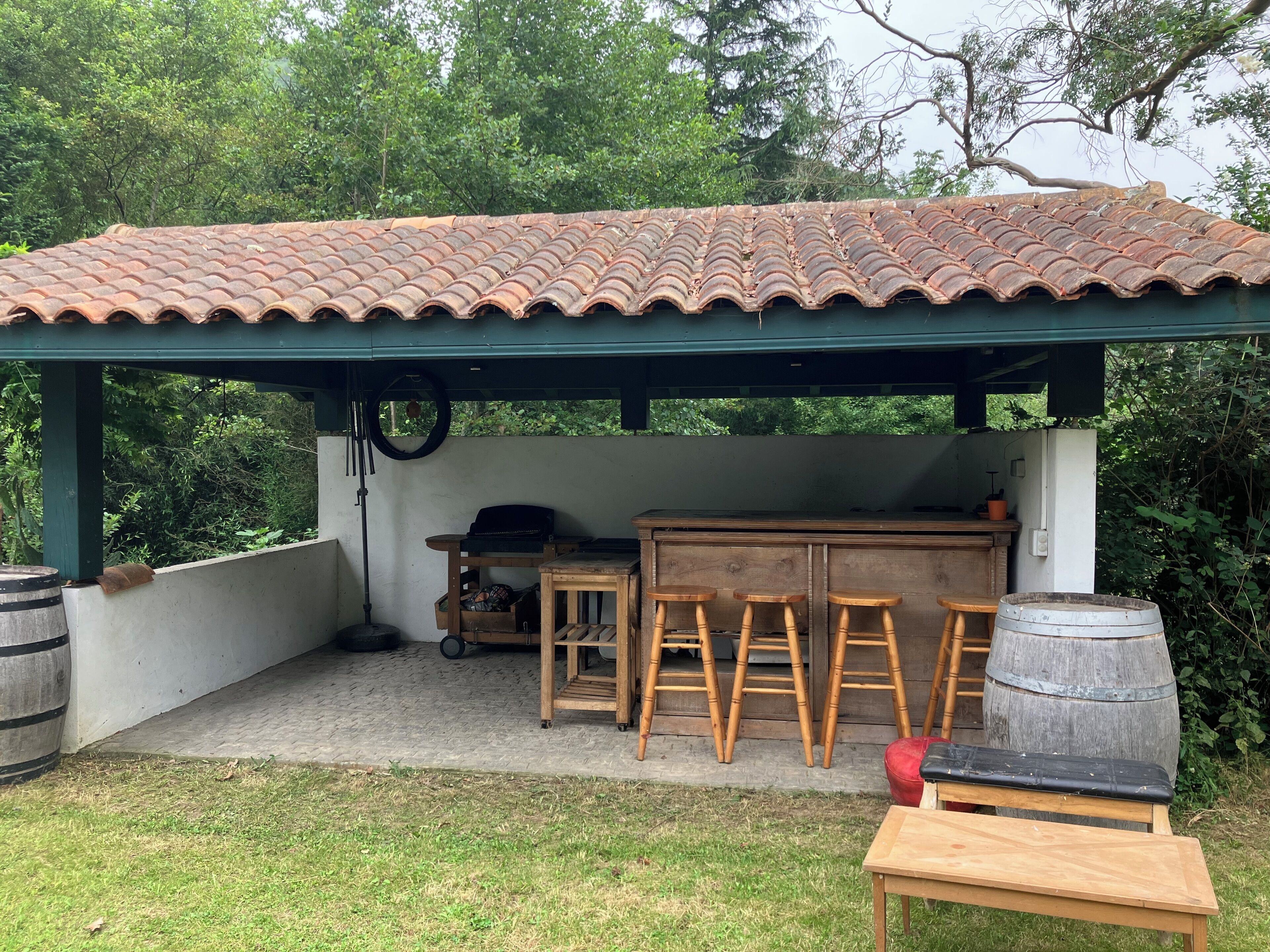 Une cuisine extérieure rustique, dotée d’un toit en tuiles, de tabourets de bar en bois, d’une table en bois et d’un tonneau.