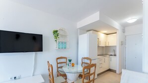 Cocina compacta con armarios blancos, un lavamanos y un refrigerador. Cuenta con una mesa redonda de vidrio con cuatro sillas, un televisor de pantalla plana montado en la pared y una pequeña planta en maceta.