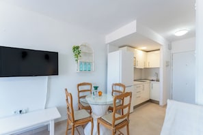 Eine kompakte Küche mit weißen Schränken, einer Spüle und einem Kühlschrank. Ein runder Glastisch wird von vier Stühlen begleitet, an der Wand ist ein Flachbildfernseher angebracht und eine kleine Topfpflanze steht dort.