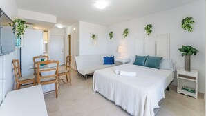 Habitación de hotel con cama, una pequeña mesa con sillas, un sofá, una mesita de noche con lámpara y plantas en la pared.