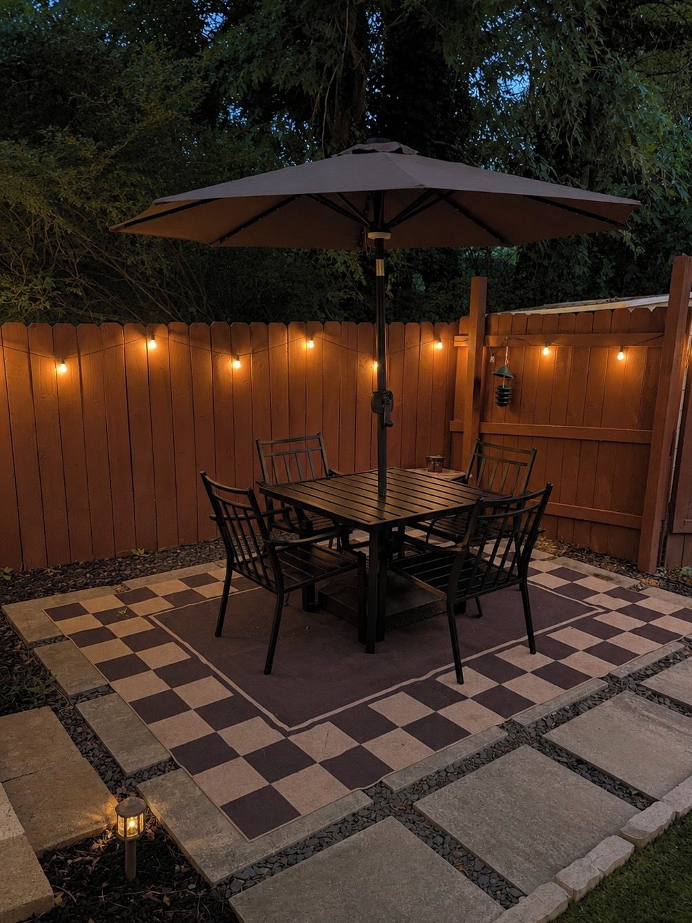 An outdoor patio with a checkered floor, a wooden table, chairs, and an umbrella.