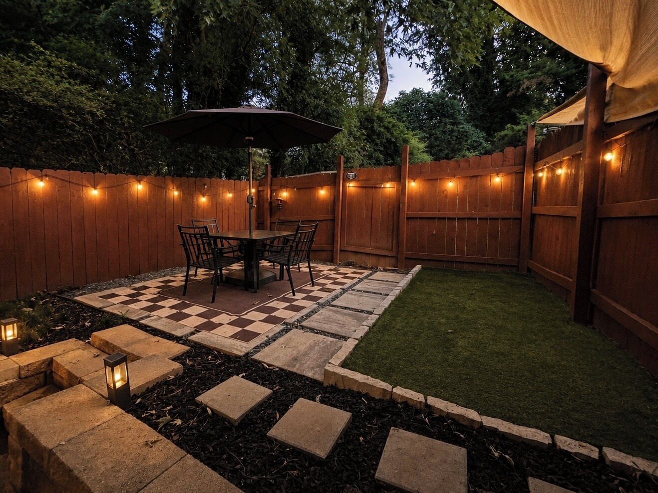 A backyard patio with a checkered stone floor, a wooden fence, and string lights.