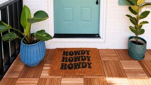 Una puerta verde azulada con un panel de vidrio, un felpudo con la inscripción 'HOWDY HOWDY HOWDY' y dos plantas en macetas sobre un porche de madera.
