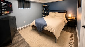 Un dormitorio con cama, cómoda, mesita de noche con lámpara, estantería y un cartel en la pared.