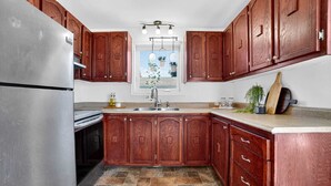 A kitchen with wooden cabinets, a stainless steel refrigerator, a window with hanging plants, and a countertop with a cutting board and a plant.