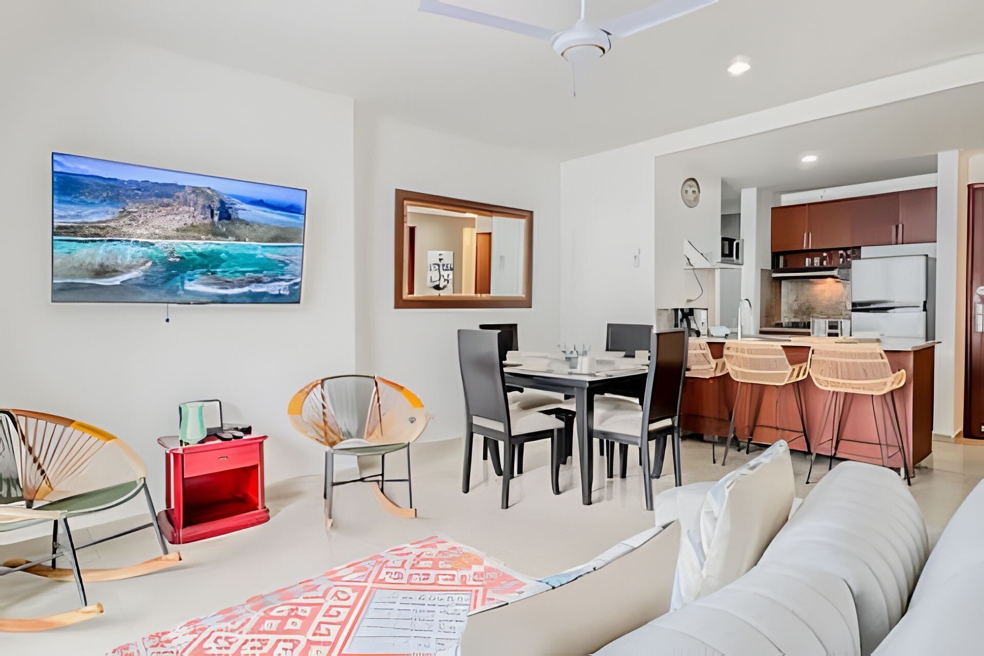 A living room with a sofa, a dining table with chairs, a kitchen area, and a large TV mounted on the wall.