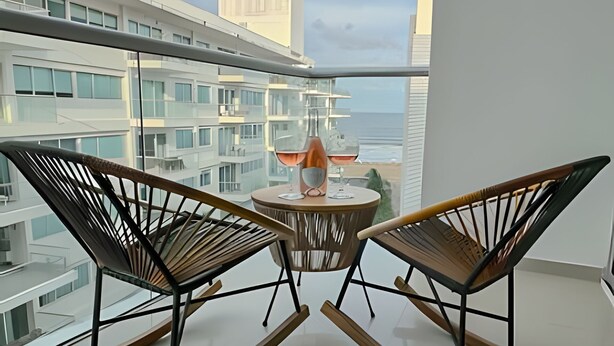 Two rocking chairs facing a balcony with a table and a glass of wine, overlooking a cityscape and the ocean. - Premium Apartment at Morros Vitri (Cartagena)