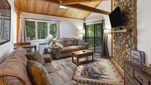 A cozy living room with a stone fireplace, a sofa, a coffee table, and a television mounted on the wall. - Escape to Ptarmigan Village 27 (Whitefish)