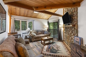 A cozy living room with a stone fireplace, a sofa, a coffee table, and a television mounted on the wall. - Escape to Ptarmigan Village 27 (Whitefish)