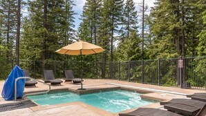 A pool area with lounge chairs and an umbrella, surrounded by a metal fence and trees. - Escape to Ptarmigan Village 27 (Whitefish)