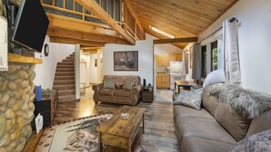 A spacious living area with a wooden ceiling, a stone wall, a sofa, a coffee table, and a staircase leading to an upper level. - Escape to Ptarmigan Village 27 (Whitefish)