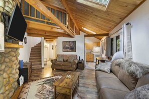 A spacious living area with a wooden ceiling, a stone wall, a sofa, a coffee table, and a staircase leading to an upper level. - Escape to Ptarmigan Village 27 (Whitefish)