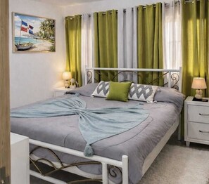Ein ordentlich bezogenes Bett mit grauer Bettdecke und Kissen, ein Gemälde mit Strandmotiv und grüne Vorhänge.