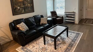 A living room with a black leather sofa, a coffee table, a patterned rug, a painting on the wall, and a door with a shoe rack beside it.