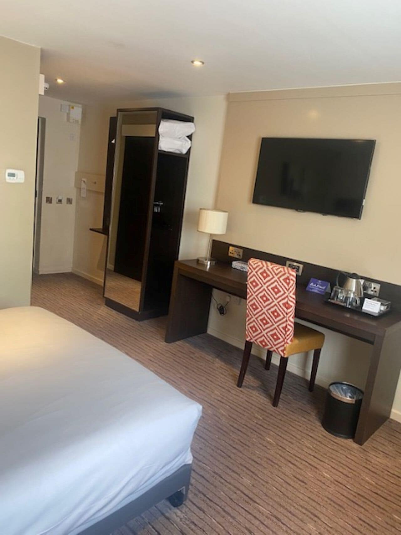 A hotel room with a bed, desk, chair, TV, and a trash bin.