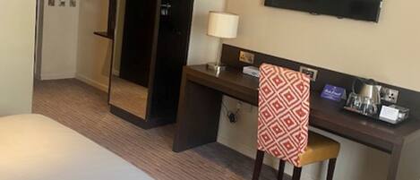 A hotel room with a bed, desk, chair, TV, and a trash bin.