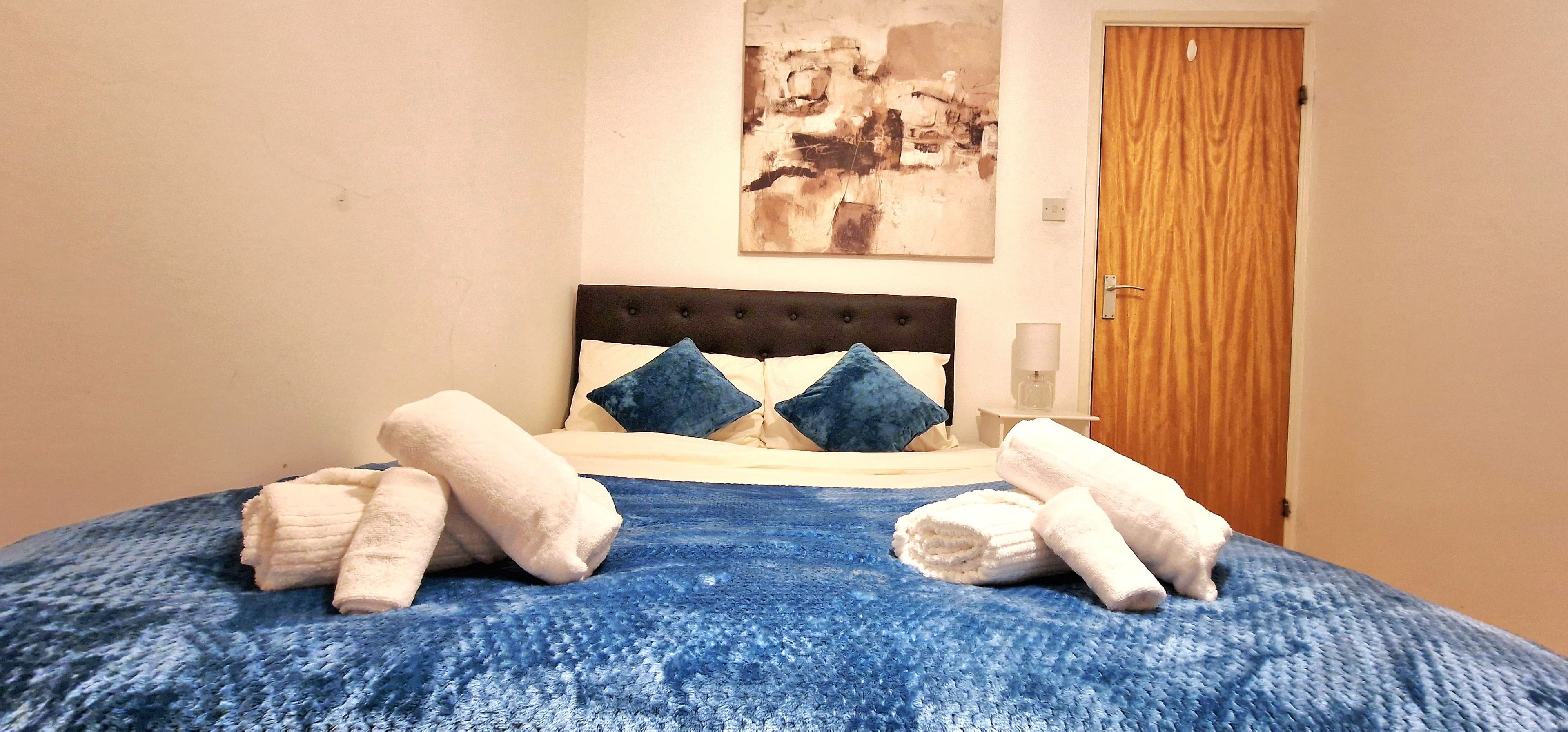Ein Bett mit blauer Bettdecke, zwei Kissen und gerollten Handtüchern.