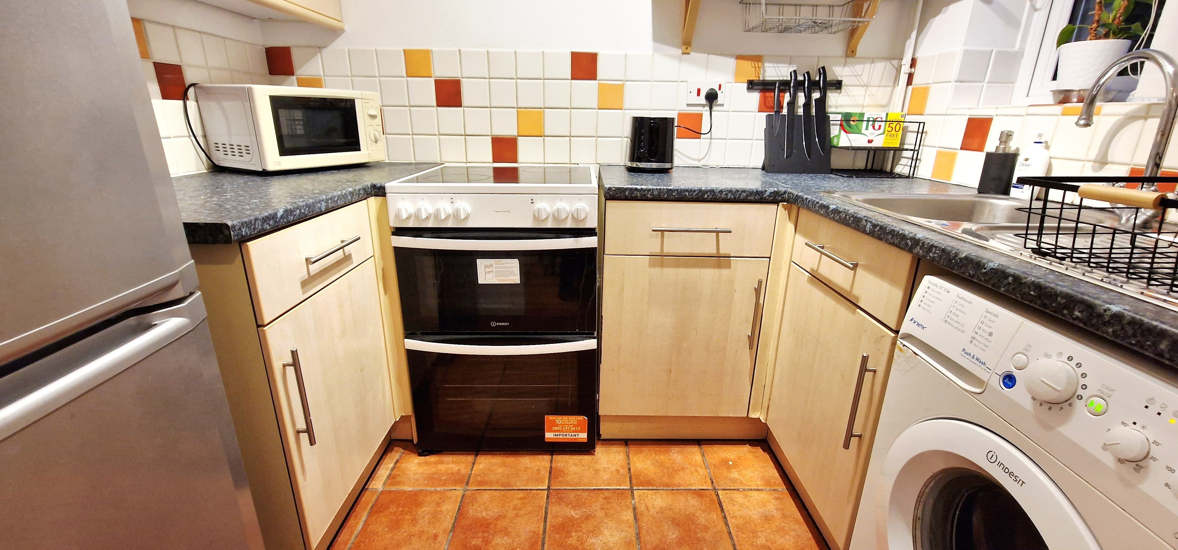 A compact kitchen with a microwave, oven, washing machine, and a tiled backsplash.