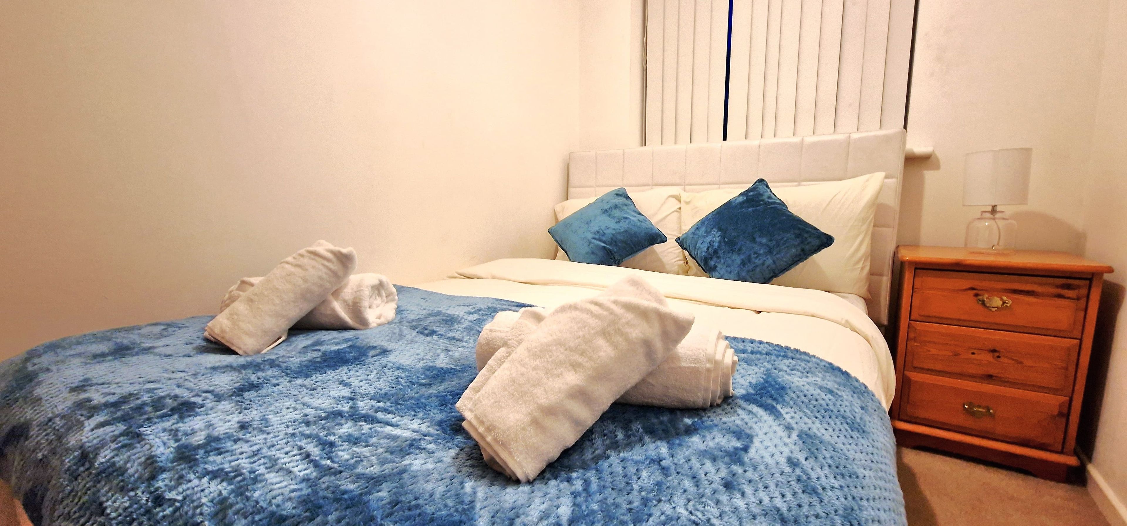Een net opgemaakt bed met een blauw-witte bedspreide, twee kussens en twee opgerolde handdoeken op het bed.