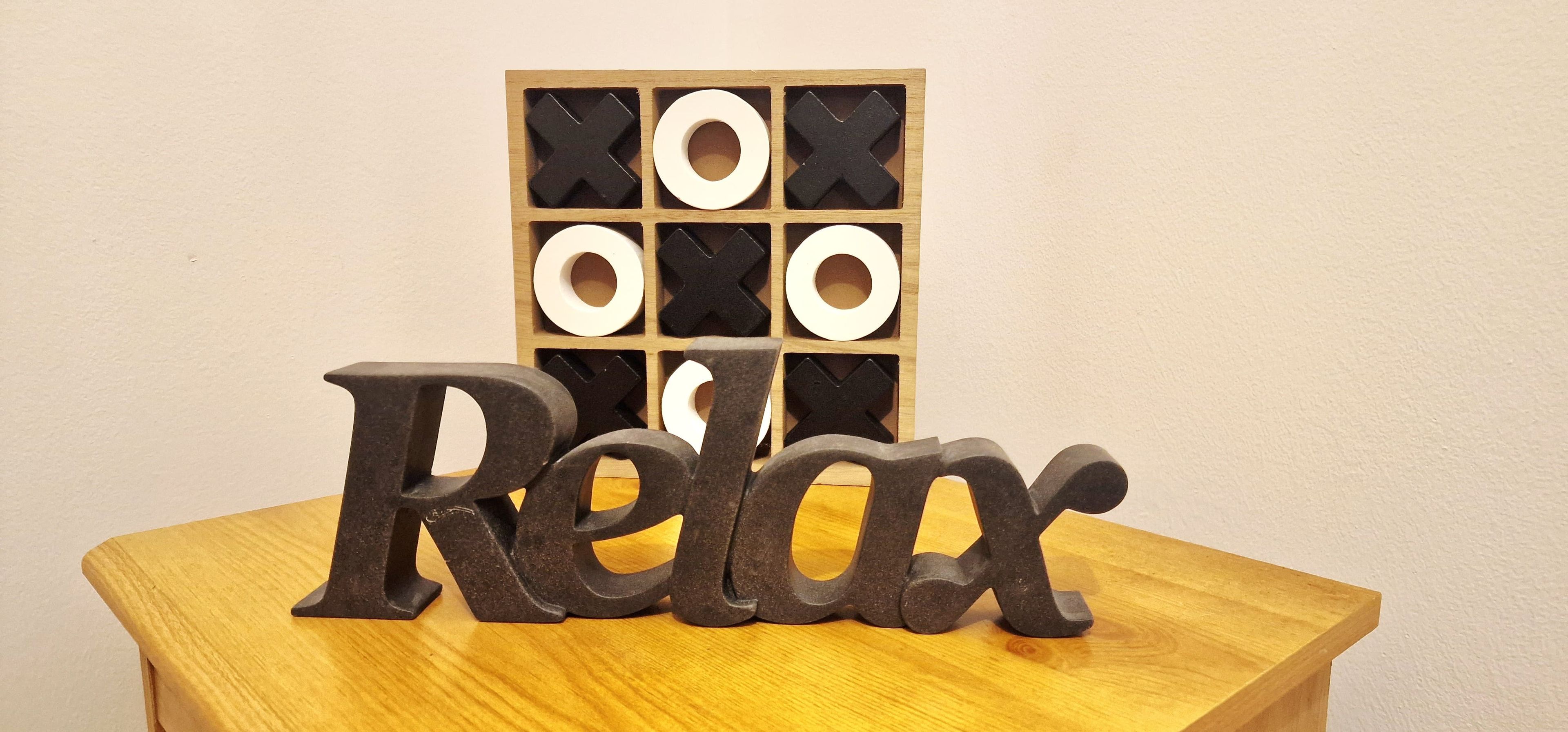 Una mesa de madera con la palabra 'Relax' en letras grandes. Detrás, un panel de madera muestra formas blancas y negras.