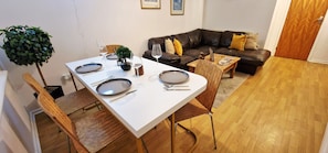 A dining area with a white table set for four, surrounded by wooden chairs. A brown leather sofa and a small wooden coffee table are in the background.