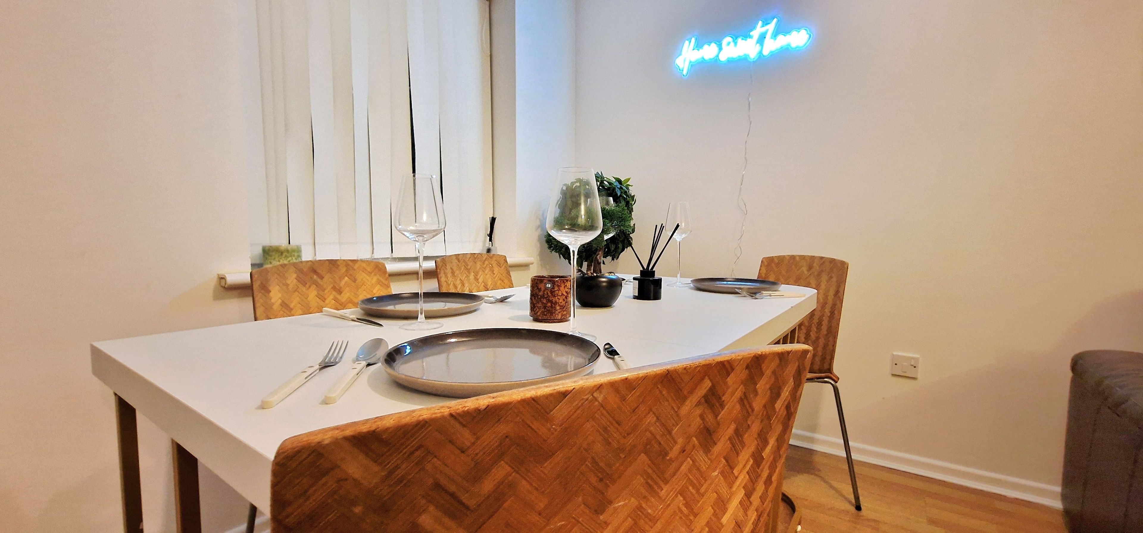 A dining area with a white table, wooden chairs, and a neon sign on the wall.