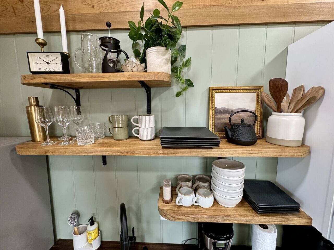 Une étagère de cuisine contenant divers objets, notamment une horloge, des verres, des tasses, une bouilloire et des ustensiles.