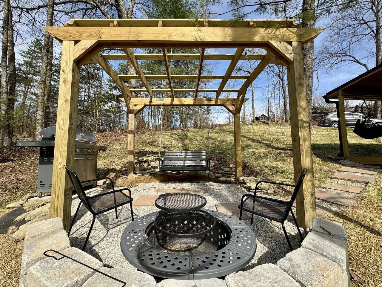 A wooden pergola structure with a fire pit in the center, surrounded by stone seating.