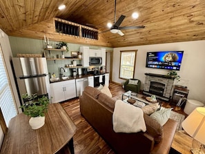 Una cocina moderna con electrodomésticos de acero inoxidable, un comedor con mesa y sillas de madera, una sala de estar con sofá y chimenea, y un televisor montado en la pared.