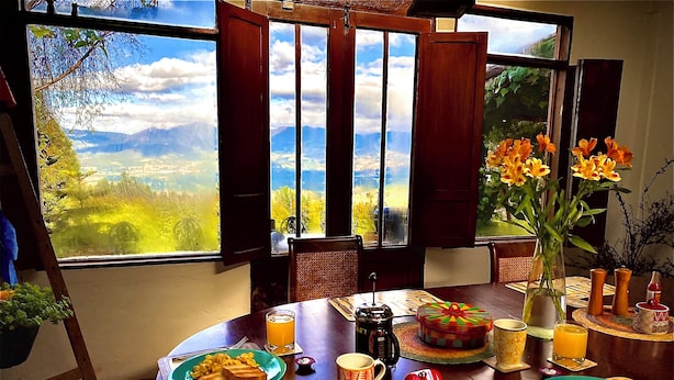 Uma mesa de jantar com itens de café da manhã, vista para montanhas e um vaso com flores.